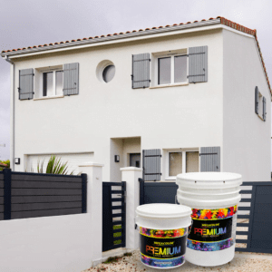 Cubetas de pintura Premium de Megacolor en primer plano, con una casa moderna de fondo pintada en un color blanco uniforme, resaltando la calidad y cobertura del látex.