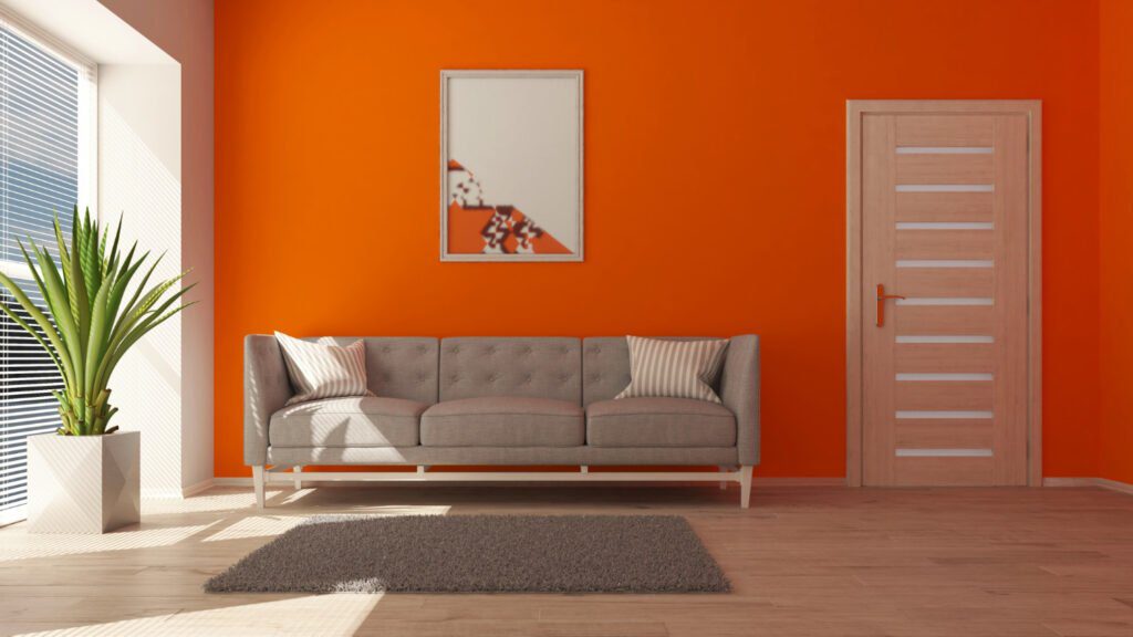 Interior moderno y elegante con paredes pintadas con la vibrante pintura naranja de Megacolor, complementando una sala de estar con un sofá gris, una alfombra gris suave, una planta verde en macetero blanco y una puerta de madera clara, demostrando cómo la pintura de Megacolor puede transformar un espacio residencial con un toque audaz y contemporáneo.