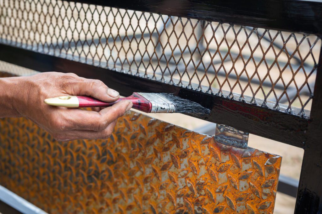 Mano experta aplicando meticulosamente pintura protectora Megacolor en una reja metálica negra sobre una textura de óxido de hojas, resaltando la calidad y durabilidad de la pintura Megacolor en condiciones exteriores, ideal para proyectos de restauración y mantenimiento metálico.