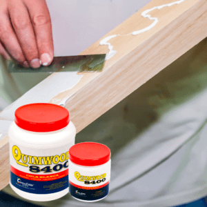 Mano aplicando cola blanca QUIMWOOD 8400 de Megacolor a una pieza de madera, con envases del producto en primer plano, destacando su uso en trabajos de carpintería y bricolaje.