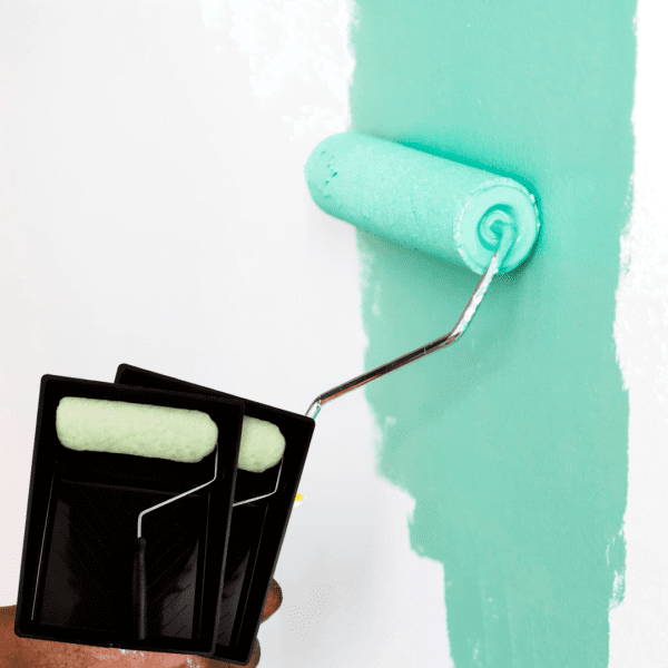 Mano aplicando pintura de color turquesa en una pared blanca con un rodillo, junto a una bandeja de pintura con otro rodillo listo para usar.