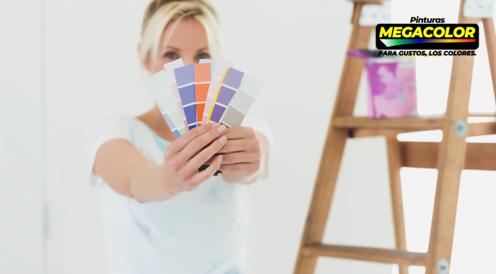 Mujer sosteniendo abanicos de muestras de colores de Pinturas Megacolor, con una escalera y una lata de pintura desenfocada al fondo, bajo el eslogan 'Para gustos, los colores'.